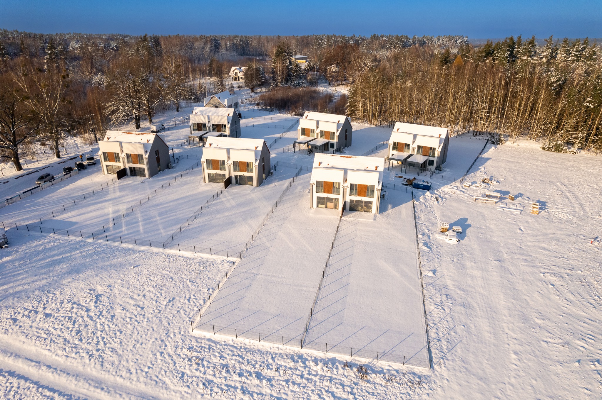 Zimowy widok z lotu ptaka na osiedle nowoczesnych domów w Olsztynie, pokrytych śniegiem, otoczonych lasem i ogrodzeniem.