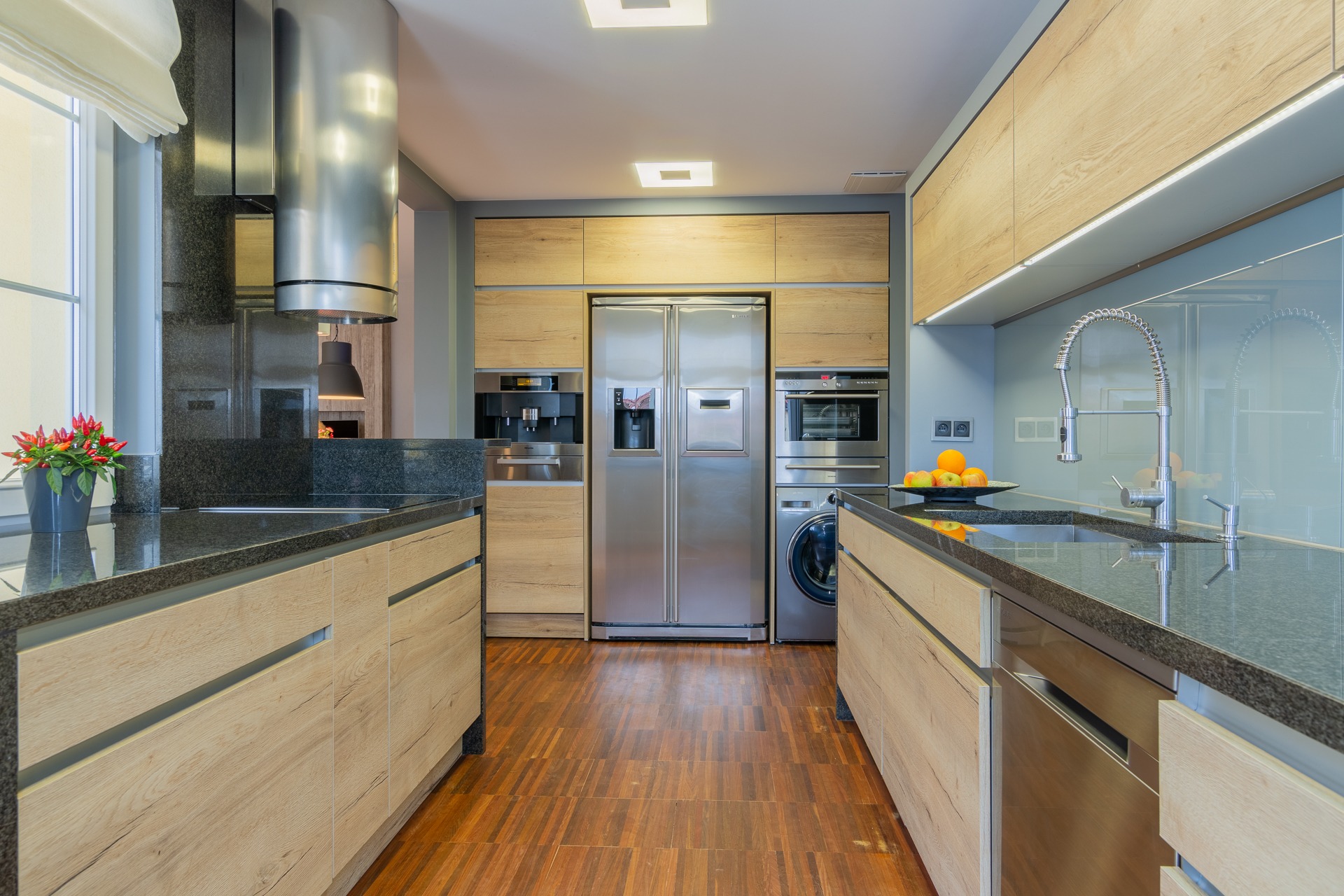 Nowoczesna kuchnia z drewnianymi szafkami, czarnym blatem, stalową lodówką i sprzętami AGD w zabudowie, oświetlona światłem z okna i lamp sufitowych.