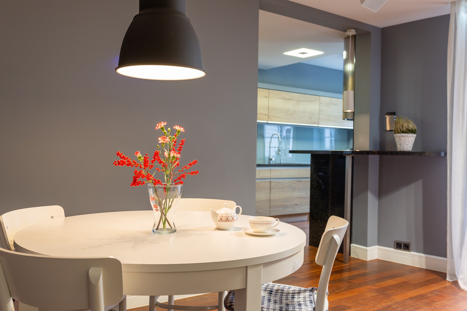 Jasny okrągły stół z bukietem czerwonych i różowych kwiatów w wazonie, serwisem do herbaty, drewniana podłoga, widok na kuchnię w tle, oświetlenie lampą wiszącą.