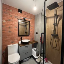 Nowoczesna łazienka z elementami cegły, czarną armaturą i kabiną prysznicową typu walk-in, szara szafka pod umywalką z drewnianym blatem, widoczna kuweta dla kota.