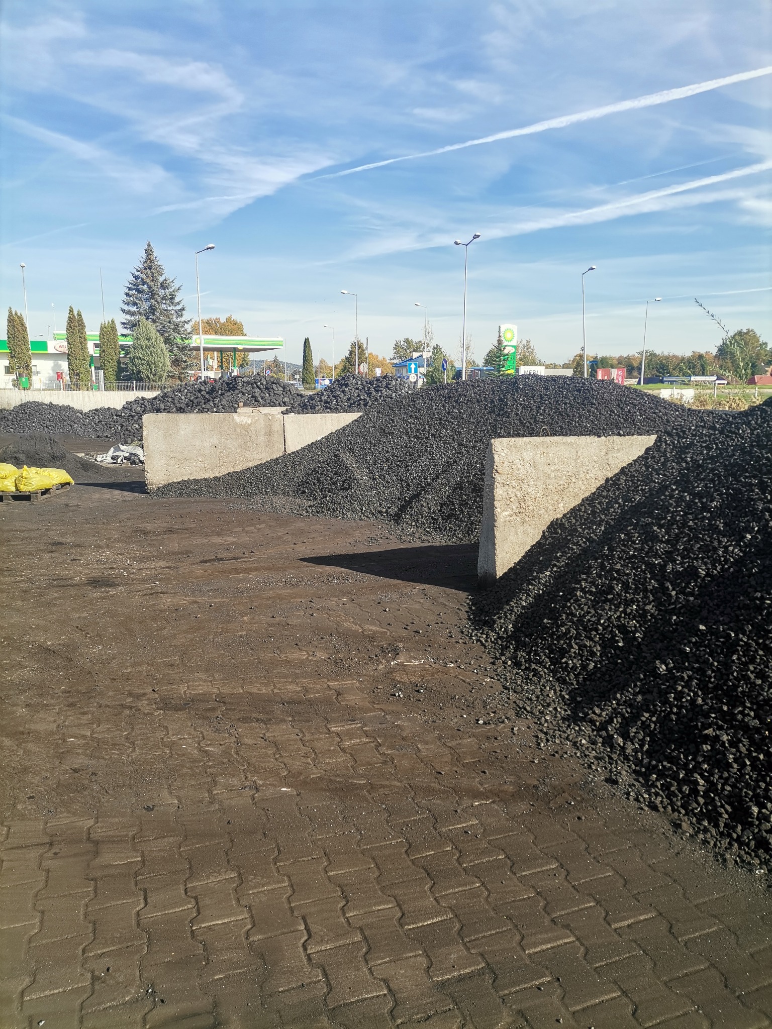 Składowisko węgla z betonowymi przegrodami, widok na hałdy czarnego węgla, żółte worki na palecie i stację benzynową w tle pod błękitnym niebem.