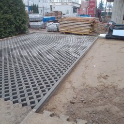 WM-bud - Częściowo ułożona szara ażurowa kostka brukowa na podbudowie z piasku, widoczne palety z materiałami budowlanymi w tle.