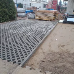 Częściowo ułożona szara ażurowa kostka brukowa na podbudowie z piasku, widoczne palety z materiałami budowlanymi w tle.