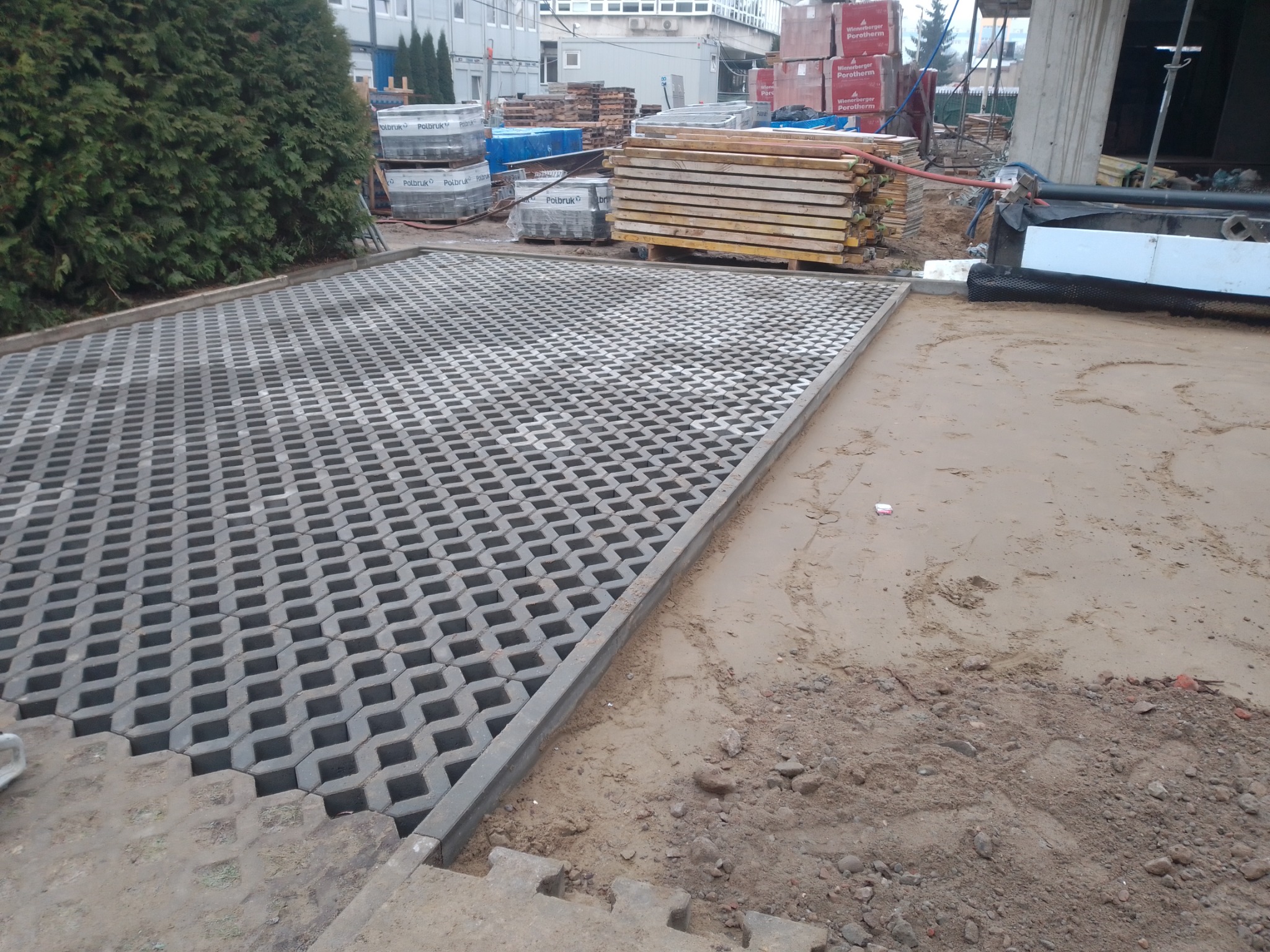 Częściowo ułożona szara ażurowa kostka brukowa na podbudowie z piasku, widoczne palety z materiałami budowlanymi w tle.