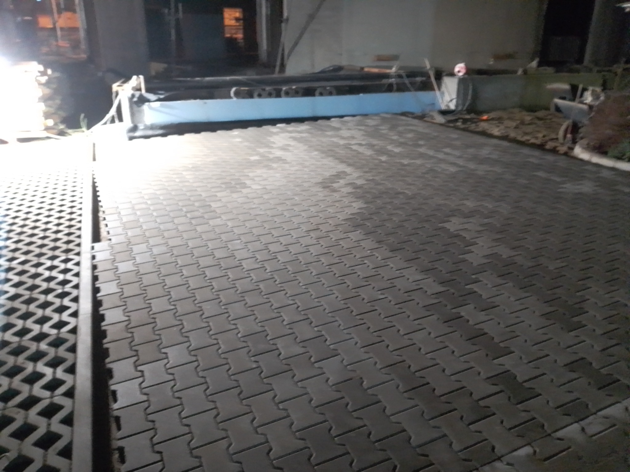 Ułożona szara kostka brukowa typu 'psią kość' na podjeździe, widoczne korytko odwadniające po lewej stronie, w tle budynek w trakcie budowy.