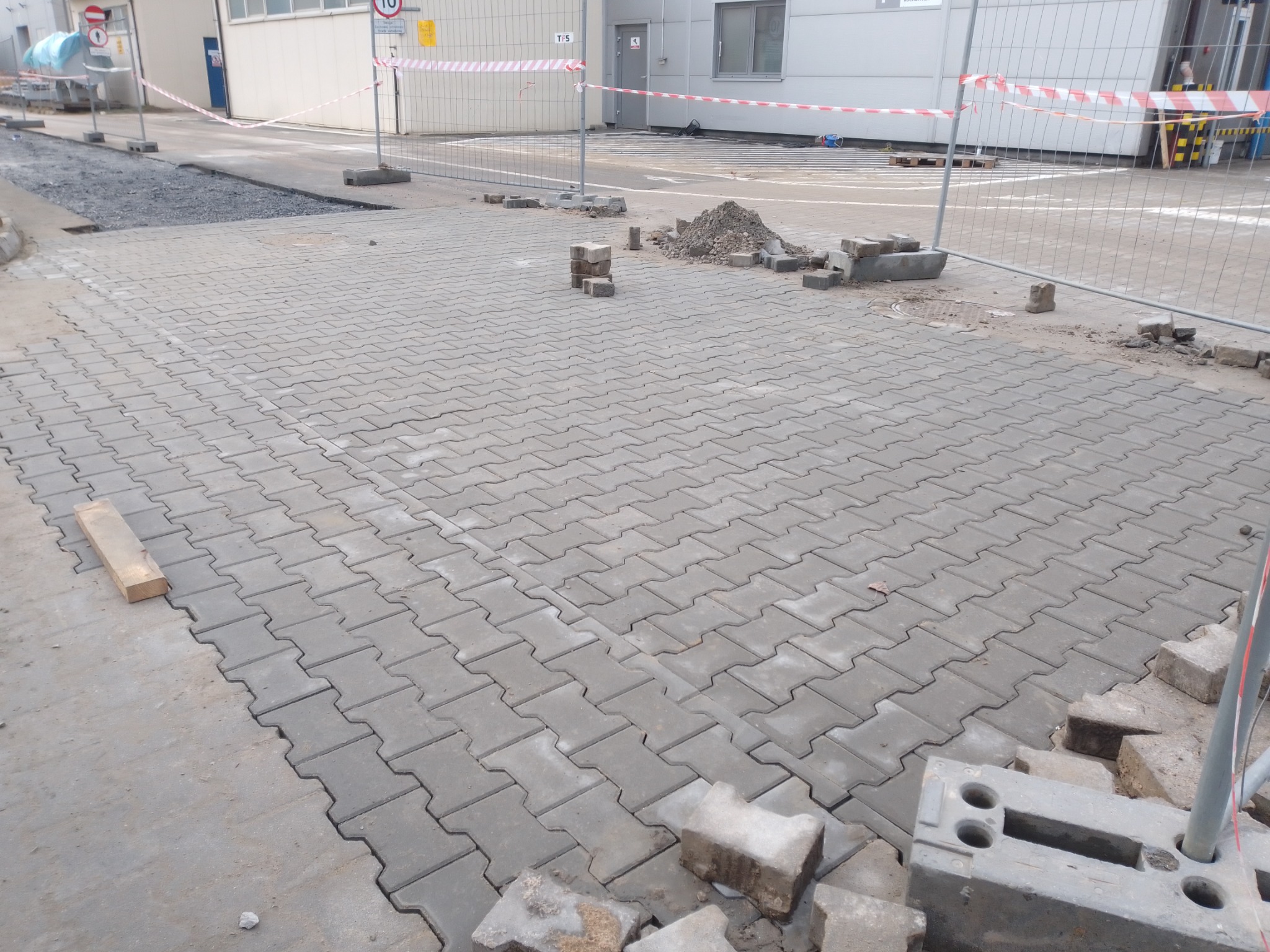 Fragment placu z ułożoną szarą kostką brukową typu 'H', w tle budynek przemysłowy i ogrodzenie z taśmą ostrzegawczą, widoczny stos kostki brukowej i niewielka pryzma piasku.