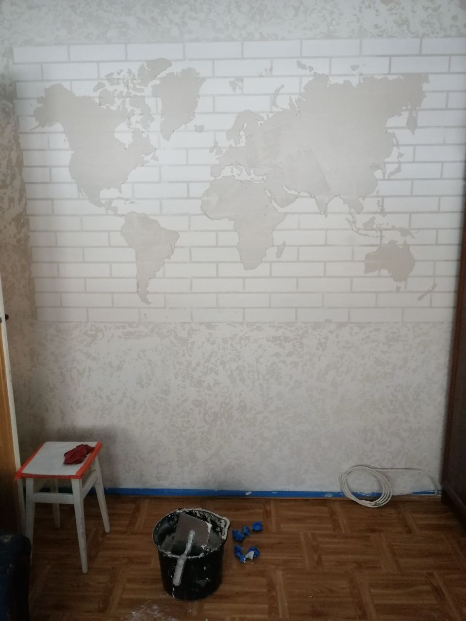 Ściana po malowaniu dekoracyjnym z efektem cegieł i mapy świata, widoczne narzędzia malarskie, wiadro z farbą, taboret i zabezpieczona podłoga.