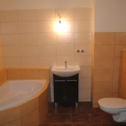 Ukończona łazienka z beżowymi płytkami, narożną wanną, umywalką na szafce i podwieszaną toaletą, oświetlona pojedynczą żarówką