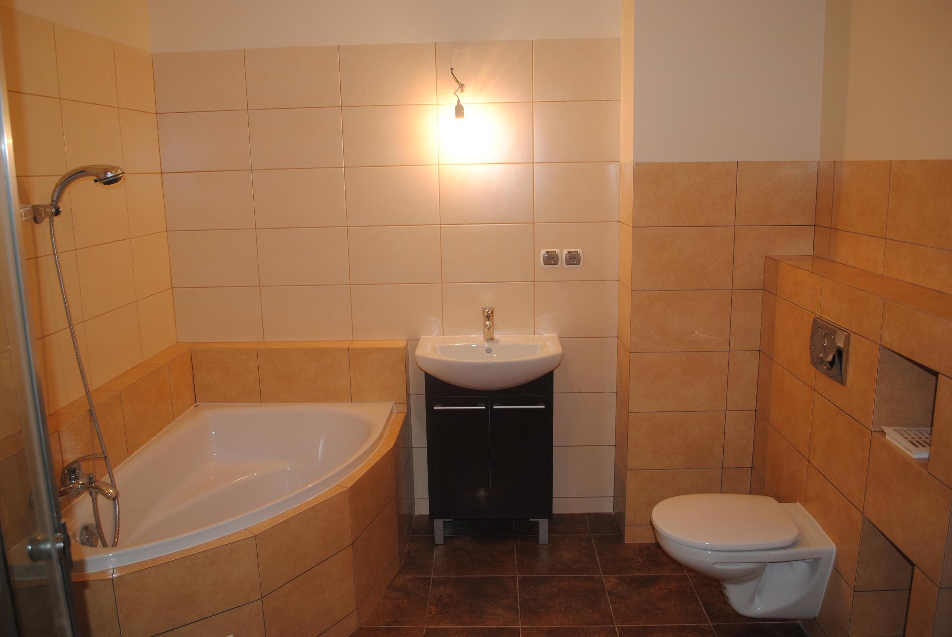 Ukończona łazienka z beżowymi płytkami, narożną wanną, umywalką na szafce i podwieszaną toaletą, oświetlona pojedynczą żarówką