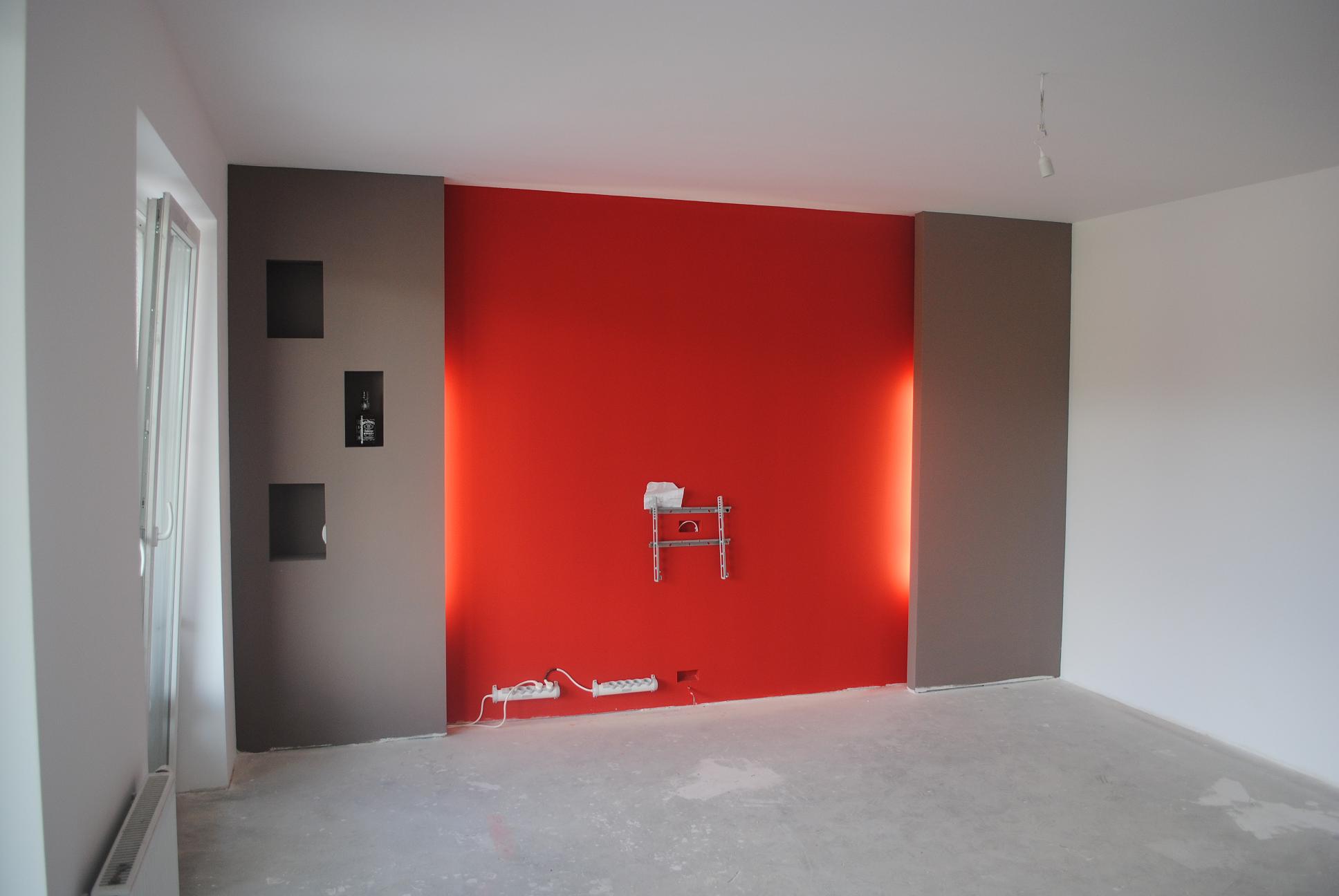 Pokój w trakcie prac wykończeniowych: czerwona ściana z uchwytem na telewizor, szare wnęki, surowa posadzka, widoczne gniazdka elektryczne i kabel od lampy sufitowej.