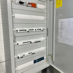 IEMS Instalacje Elektryczne Michał Sulich - Otwarta szafa elektryczna z widocznymi bezpiecznikami, wyłącznikami i przewodami, żółta naklejka ostrzegawcza na wewnętrznej stronie drzwi.