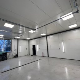 IEMS Instalacje Elektryczne Michał Sulich - Wnętrze hali z panelami ściennymi i oświetleniem LED, widoczny drenaż podłogowy, okno z widokiem na drzewa i elementy wyposażenia budowlanego.