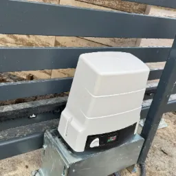 Szary metalowy płot z zamontowanym białym napędem do bramy z widoczną listwą zębatą i logo Roger Technology, na lekko zabrudzonym podłożu.