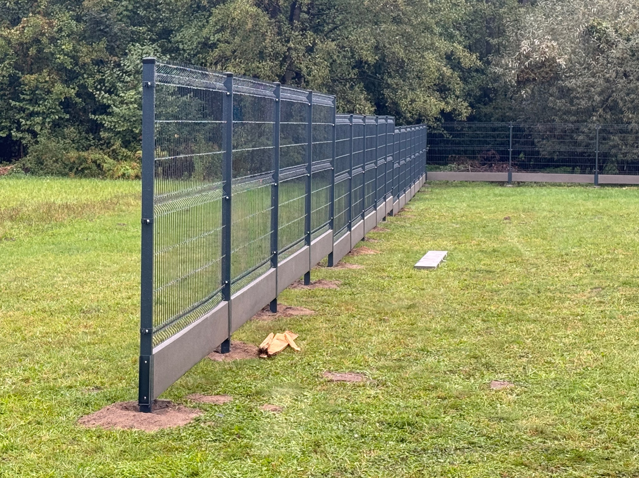 Długa, szara siatka ogrodzeniowa panelowa z betonową podmurówką, biegnąca wzdłuż zielonego trawnika, widok z boku, z liśćmi i gałęziami drzew w tle.