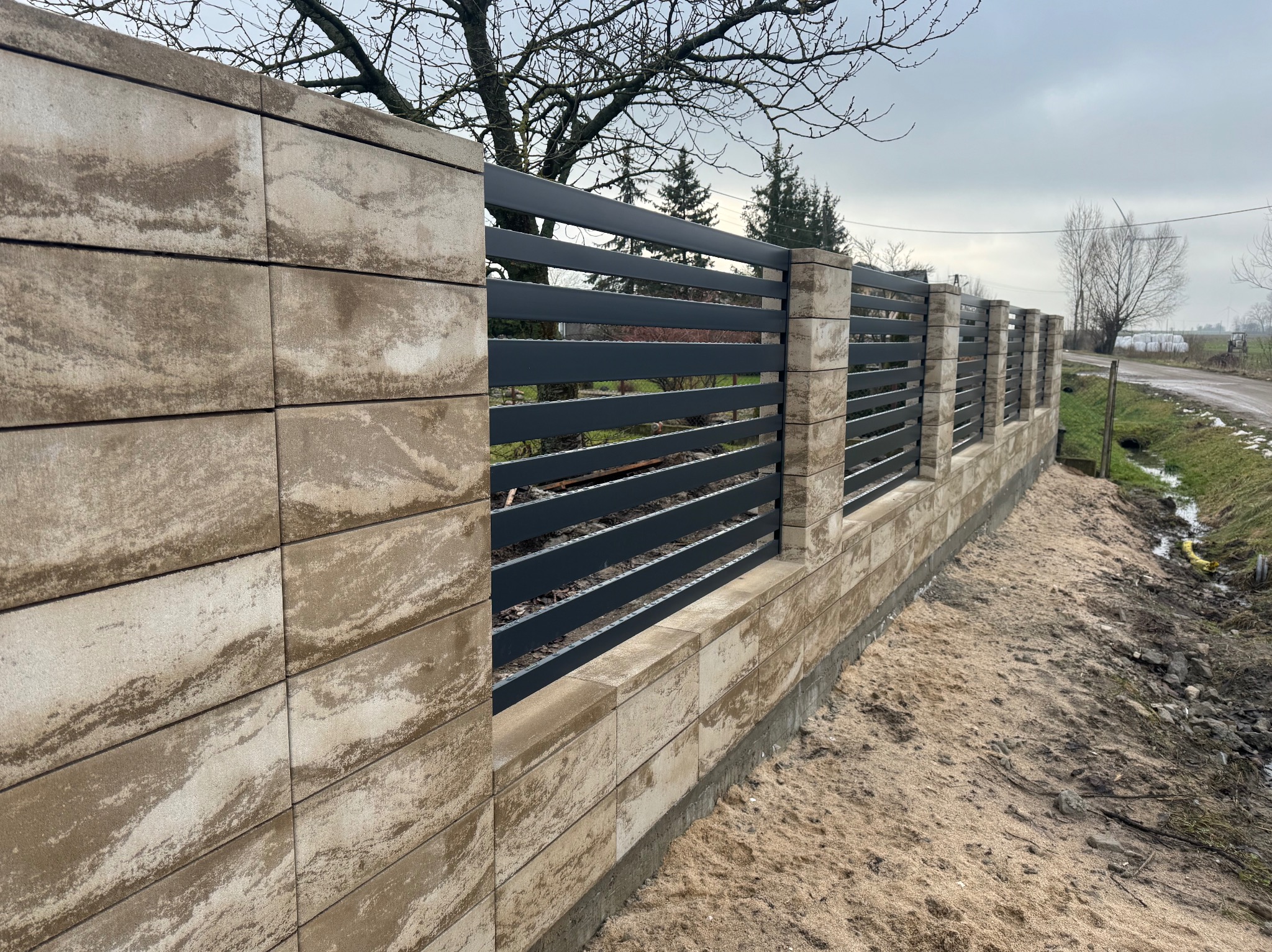 Nowoczesne ogrodzenie z poziomych, grafitowych paneli między słupkami z beżowych, teksturowanych bloczków betonowych, widoczne ślady świeżego montażu na podłożu.