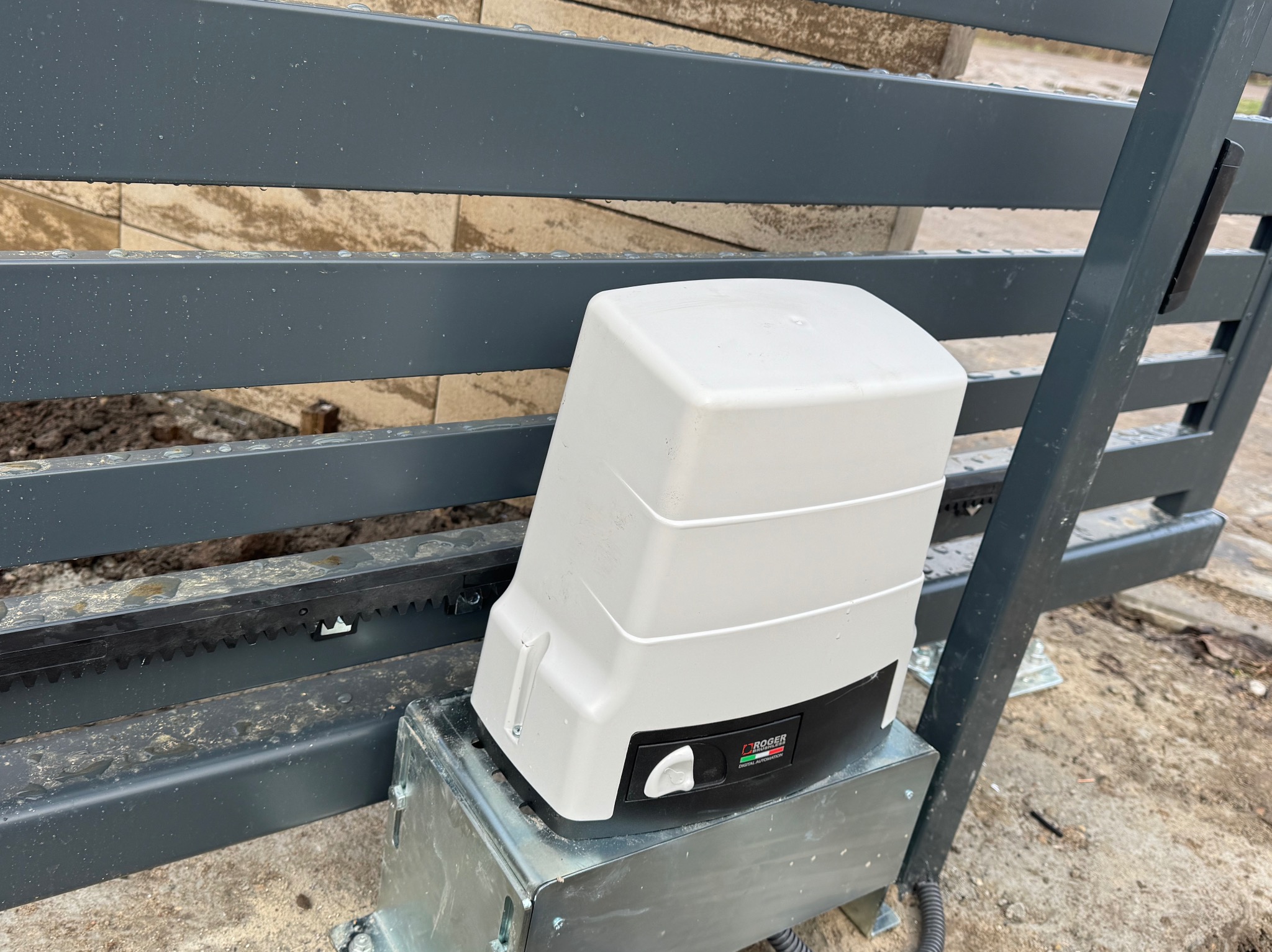 Szary metalowy płot z zamontowanym białym napędem do bramy z widoczną listwą zębatą i logo Roger Technology, na lekko zabrudzonym podłożu.