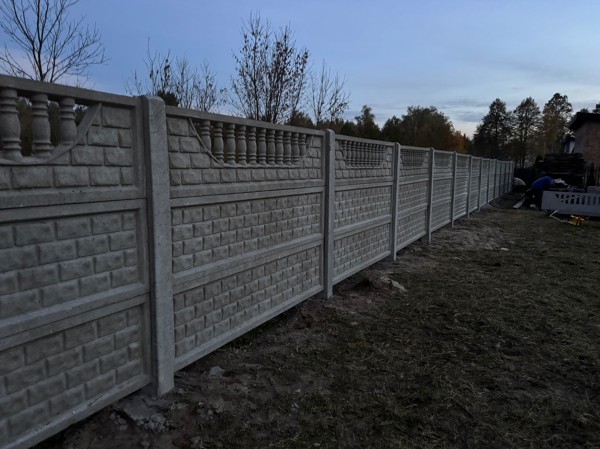 Długi, szary, betonowy płot panelowy z dekoracyjnym wzorem cegieł i balustrad na górze, w tle drzewa i błękitne niebo o zmierzchu, widoczny fragment budynku i pracownik w oddali.