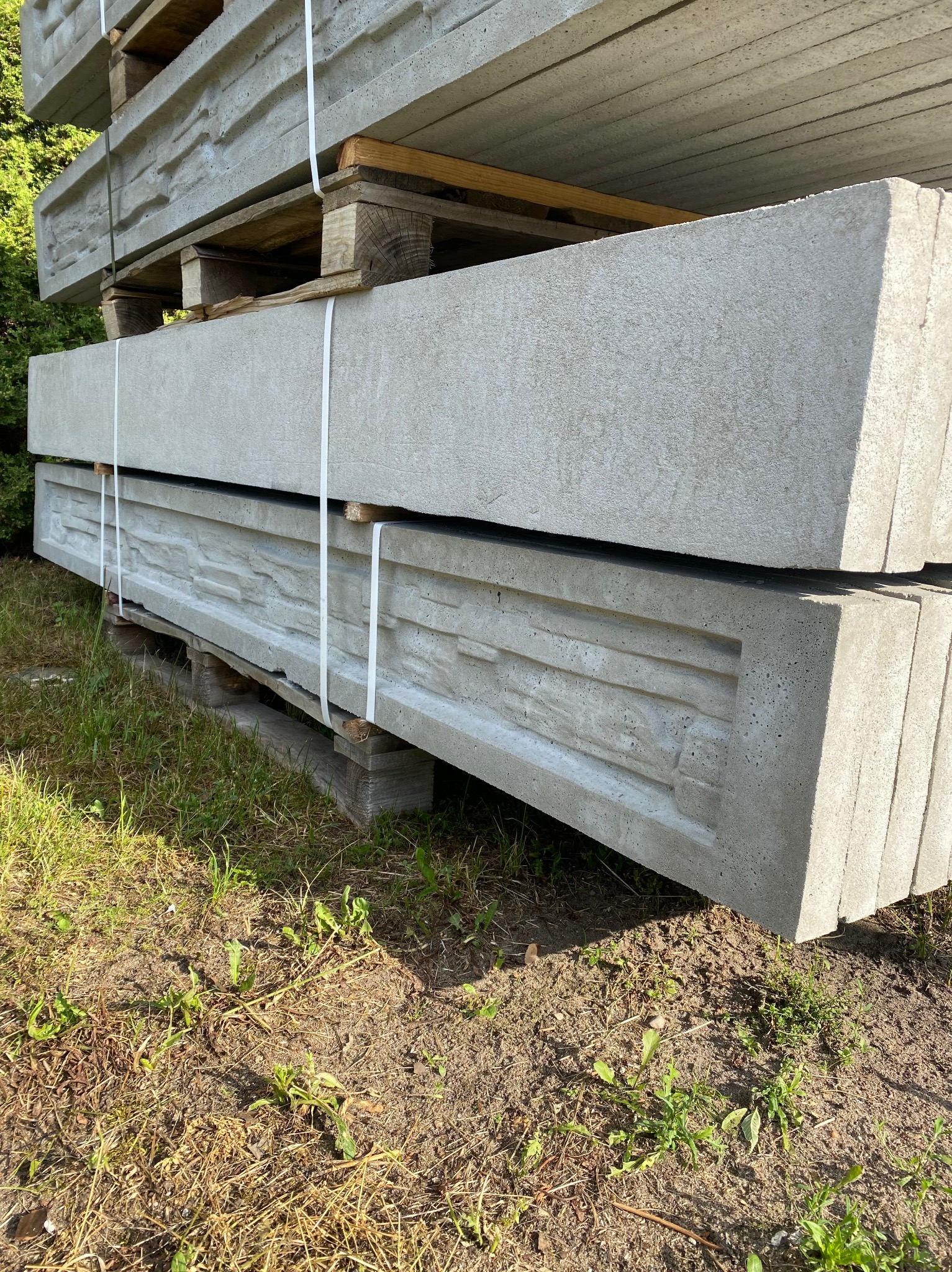 Paleta z betonowymi podmurówkami ogrodzeniowymi z dekoracyjnym wzorem kamienia, zabezpieczona białymi taśmami, leżąca na trawie przy zieleni.