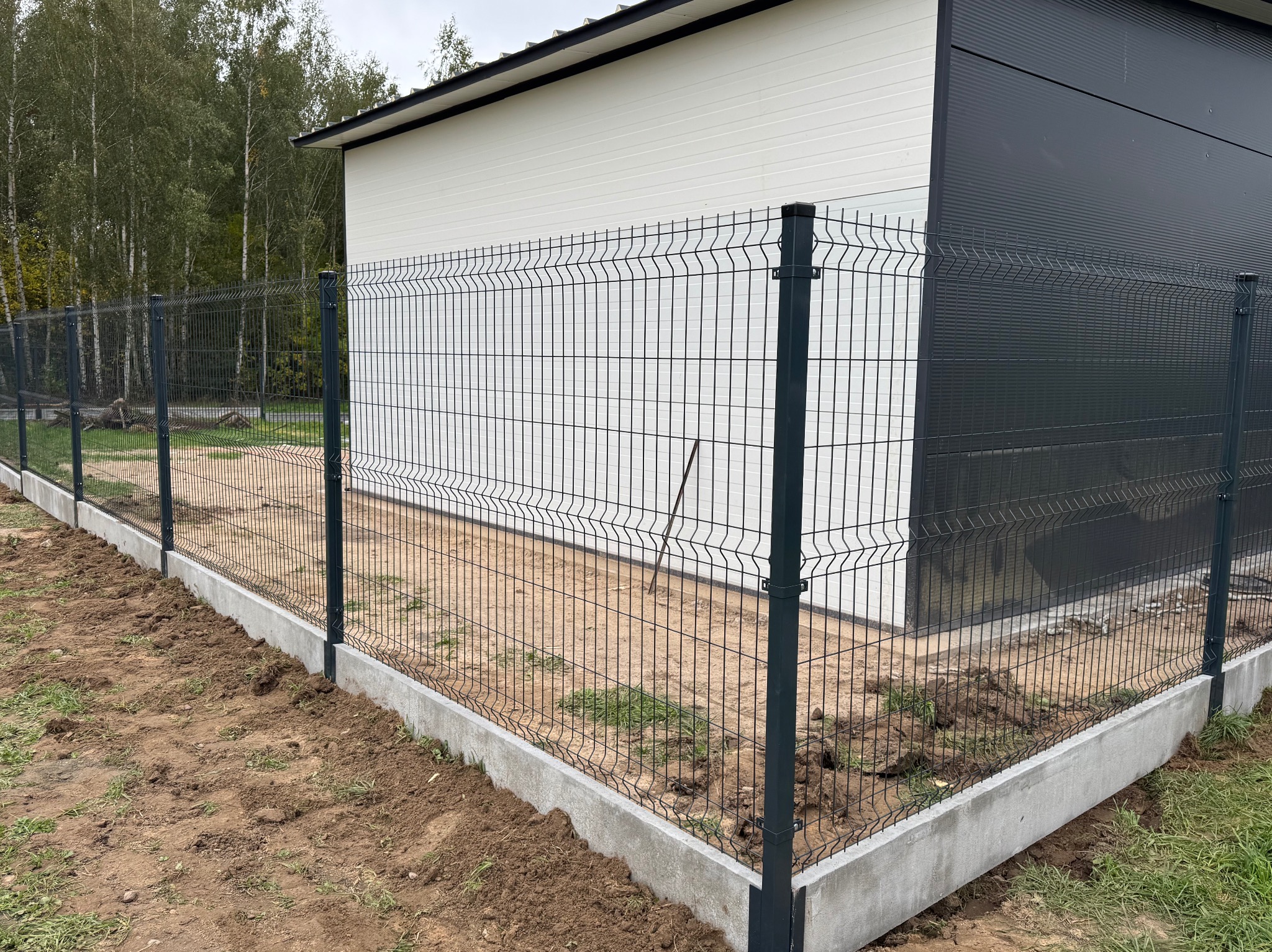 Nowo zamontowane ogrodzenie panelowe z podmurówką betonową przylegające do budynku z elewacją w kolorze białym i grafitowym, widoczne ślady prac ziemnych.