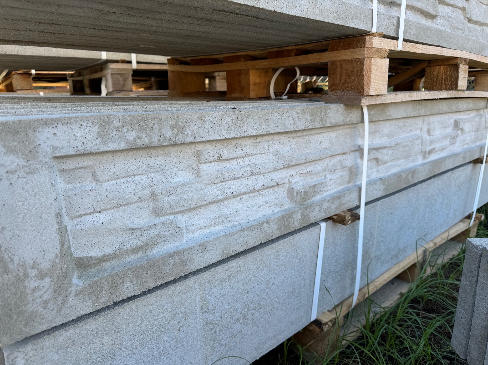 Stos prefabrykowanych, betonowych elementów ogrodzeniowych z dekoracyjnym wzorem imitującym kamień, spiętych taśmami na drewnianych paletach, widoczne źdźbła trawy w tle.
