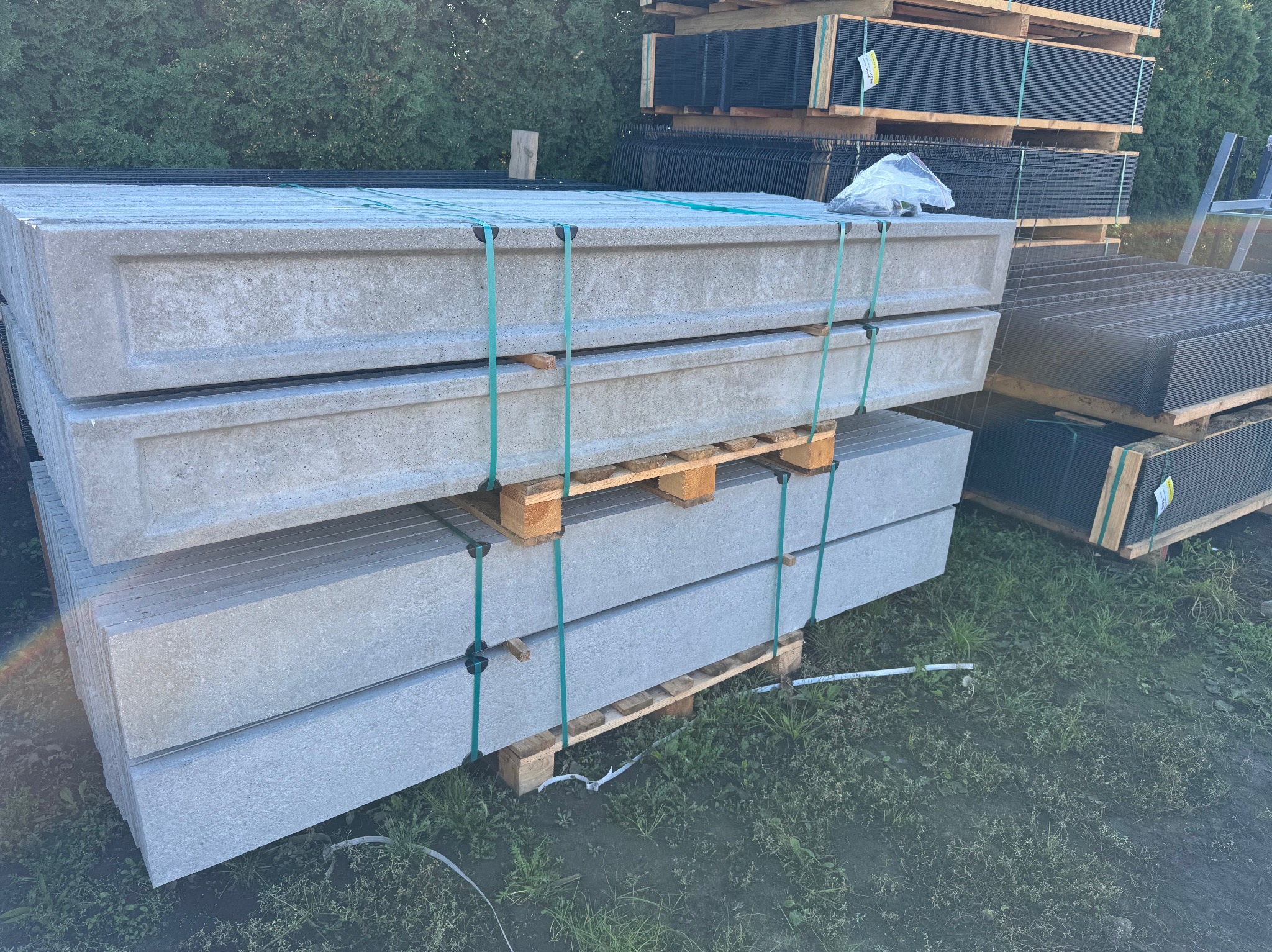 Stos prefabrykowanych, betonowych podmurówek ogrodzeniowych na paletach, zabezpieczonych zielonymi taśmami, obok składowane panele ogrodzeniowe.