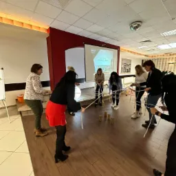 Grupa osób uczestniczy w grze zespołowej z wykorzystaniem sznurków i drewnianych elementów, stojąc w kole na drewnianej podłodze, w tle biała ściana z czerwonym akcentem i ekran projektora.