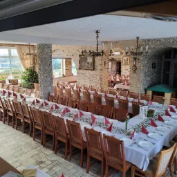 Sala bankietowa z długimi stołami nakrytymi białymi obrusami, czerwonymi serwetkami i zielonymi dekoracjami, przygotowana na przyjęcie, z widoczną choinką w tle.