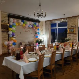 Uroczysta sala z długim stołem nakrytym białym obrusem, dekoracje z balonów i kwiatów, krzesła z drewnianymi oparciami, napoje w szklanych karafkach i butelkach, świeczniki ze świecami.