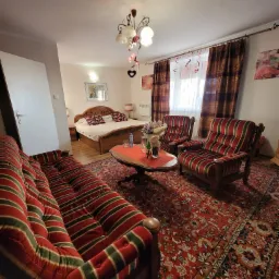 Pokój hotelowy z łóżkiem małżeńskim, sofą i fotelami w stylu retro, udekorowany na romantyczny pobyt, z bukietem kwiatów na okrągłym stoliku i girlandami w kształcie serc przy oknie.