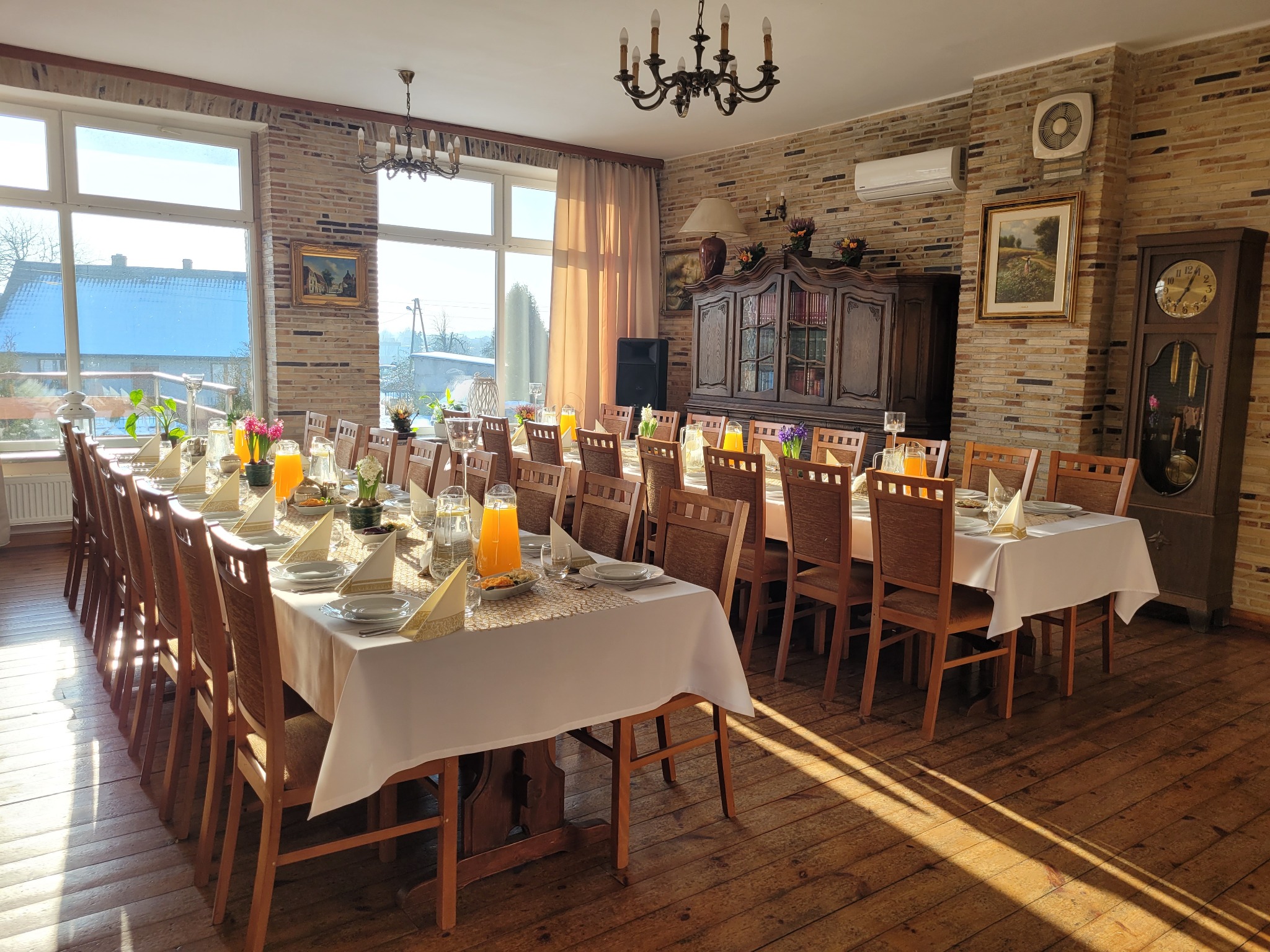 Uroczysta sala z długimi stołami nakrytymi białymi obrusami, zastawionymi talerzami, szklankami z sokiem pomarańczowym i dekoracjami kwiatowymi, oświetlona naturalnym światłem wpadającym przez duże...