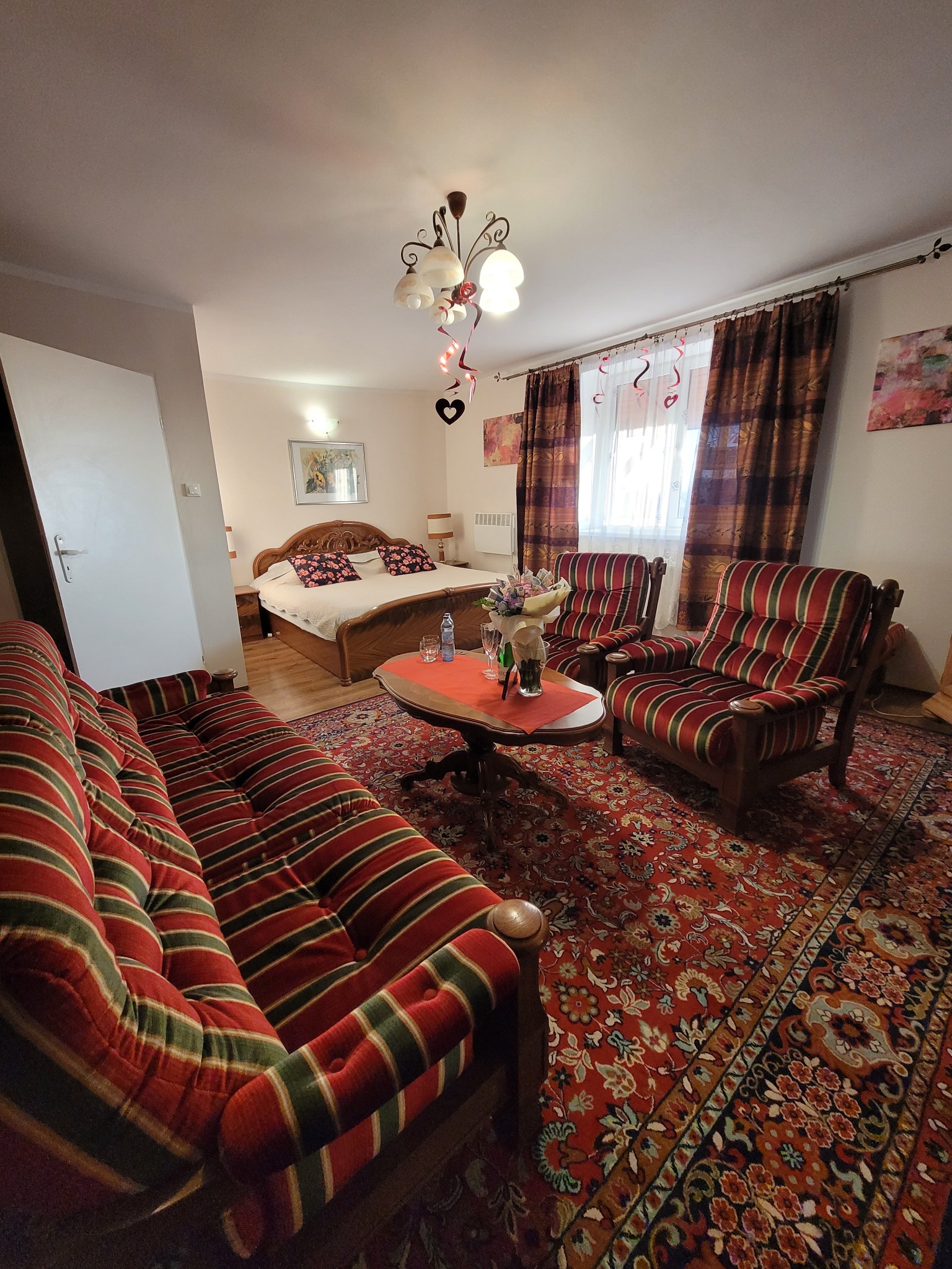 Pokój hotelowy z łóżkiem małżeńskim, sofą i fotelami w stylu retro, udekorowany na romantyczny pobyt, z bukietem kwiatów na okrągłym stoliku i girlandami w kształcie serc przy oknie.