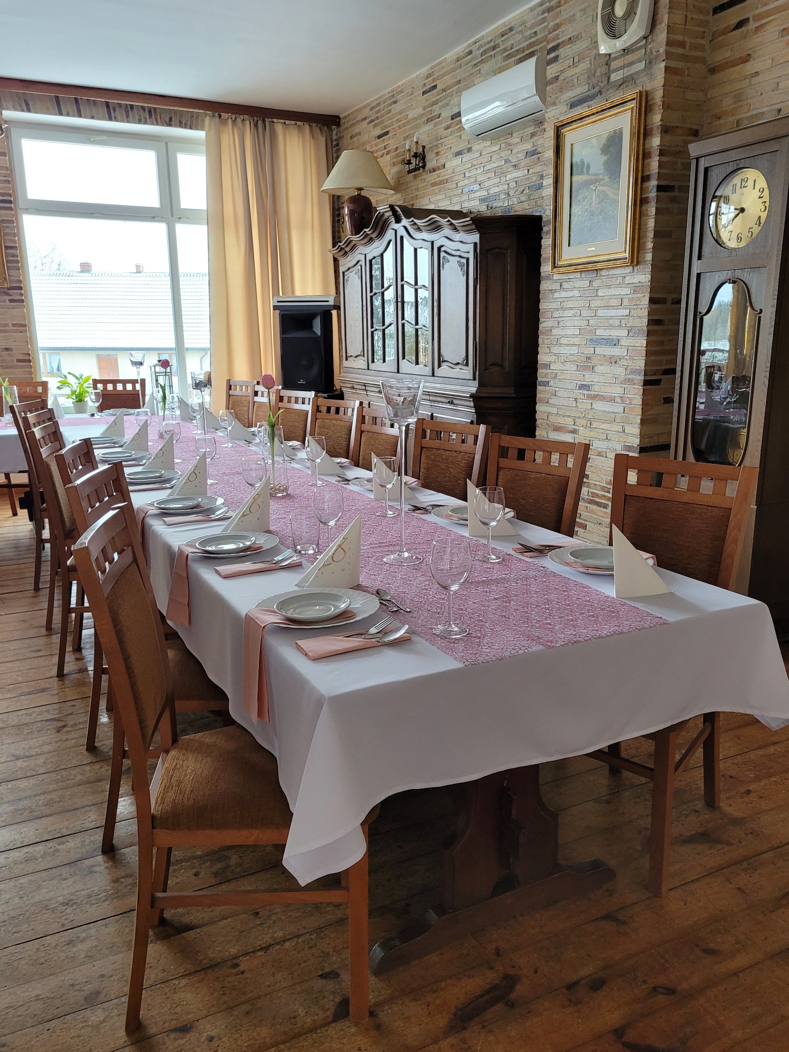 Długi stół z białym obrusem, różowym bieżnikiem, nakrytymi talerzami, sztućcami, serwetkami i kieliszkami, przygotowany do uroczystości w rustykalnym wnętrzu z drewnianą podłogą i ceglaną ścianą.