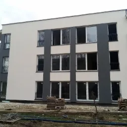 Nowo wybudowany, trzypiętrowy budynek mieszkalny z balkonami i oknami w białych ramach, w trakcie wykończenia, z paletami na ziemi i widocznymi rurami.