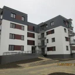 Nowoczesny budynek wielorodzinny z balkonami, wykończony w kolorach biało-szarych z brązowymi akcentami, widoczny podjazd dla niepełnosprawnych oraz częściowo zaparkowany samochód.