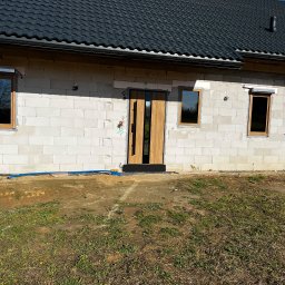 PRO MATYLDA GRAJKO - Surowy stan budynku z zamontowanymi oknami z roletami i drzwiami zewnętrznymi z drewnianym wykończeniem. Widoczne bloczki betonowe i czarna dachówka.