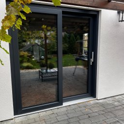 PRO MATYLDA GRAJKO - Nowoczesne, przesuwne drzwi tarasowe w grafitowej ramie, z widokiem na ogród i szklarnię. Pergola z winoroślą nad wejściem. Eleganckie wykończenie.