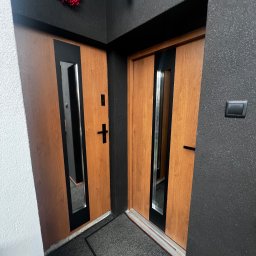 PRO MATYLDA GRAJKO - Dwa drewniane skrzydła drzwiowe z czarnymi wstawkami i lustrami, widoczne od strony wewnętrznej, z dekoracyjnym wieńcem nad jednym z nich. Ciemna ściana z fakturą.