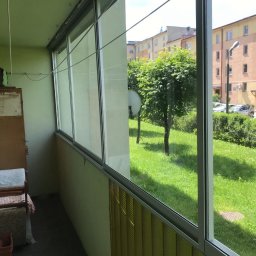 PRO MATYLDA GRAJKO - Zabudowany balkon z przesuwnymi oknami aluminiowymi, widok na zieleń. Żółte żaluzje pionowe w dolnej części okien. Widoczne budynki w tle.