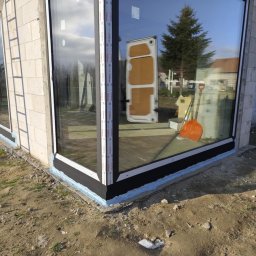 PRO MATYLDA GRAJKO - Narożne okno z czarną ramą w surowym budynku, widoczna izolacja, drabina oparta o ścianę. W odbiciu szyby widoczny samochód dostawczy i drzewo.