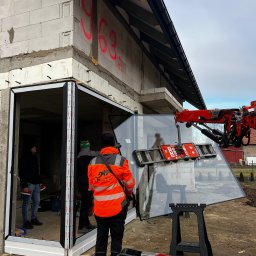 PRO MATYLDA GRAJKO - Montaż okna z użyciem dźwigu przyssawkowego na budowie. Widoczni pracownicy w odblaskowych ubraniach. Surowy stan budynku z białego bloczka.