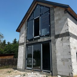 PRO MATYLDA GRAJKO - Dom w trakcie budowy z zamontowanymi oknami aluminiowymi o dużych przeszkleniach. Widoczne białe bloczki konstrukcyjne i gruz budowlany na ziemi.