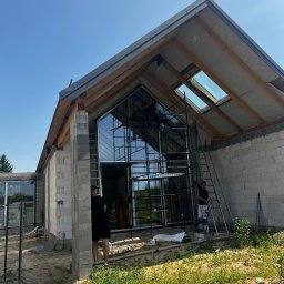 PRO MATYLDA GRAJKO - Montaż okien z czarnymi szprosami w surowym budynku z betonu komórkowego. Pracownicy na rusztowaniu i drabinie. Widoczne okno dachowe i elementy konstrukcji dachu.