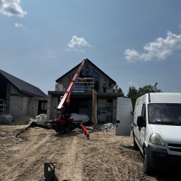 PRO MATYLDA GRAJKO - Montaż okien na budowie domu jednorodzinnego z użyciem dźwigu. Widoczny biały van i ekipa montażowa na wysokościach. Dom w stanie surowym.