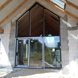 PRO MATYLDA GRAJKO - Montaż okien aluminiowych w surowym budynku. Widoczne duże przeszklenia z czarnymi ramami i drewniana konstrukcja dachu. W tle zieleń.