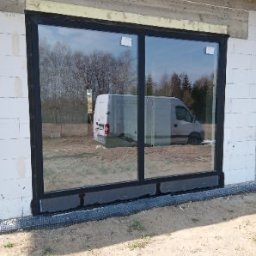 PRO MATYLDA GRAJKO - Nowe, duże okno przesuwne z czarną ramą, zamontowane w surowym murze z betonu komórkowego. Widoczne zabezpieczenia montażowe i odbicie samochodu w szybie.