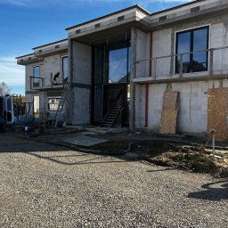 PRO MATYLDA GRAJKO - Budowa domu jednorodzinnego w stanie surowym otwartym. Widoczne okna, pustaki, rusztowania i pracownik na drabinie. Słoneczny dzień, błękitne niebo.