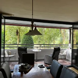 Otwarta przestrzeń salonu przechodząca w balkon z drewnianą podłogą, widok na zieleń drzew przez duże rozsuwane okna, w tle meble ogrodowe i dekoracje.