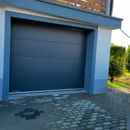 Nowoczesna, segmentowa brama garażowa w kolorze grafitowym, z poziomymi przetłoczeniami, zamontowana w szarej elewacji domu z drewnianą konstrukcją nad wjazdem, widoczna z zewnątrz na tle kostki...