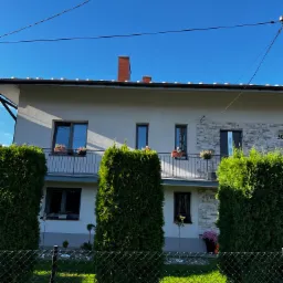 Dwupiętrowy dom z szarymi oknami i balkonem z kwiatami, fasada częściowo wykończona kamieniem, otoczony zielonymi krzewami i ogrodzeniem z siatki.