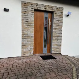 Nowoczesne drzwi wejściowe z drewnianym panelem i szarym obramowaniem, otoczone ceglaną ramą na tle białej ściany budynku, widoczne z zewnątrz na brukowanym podjeździe.